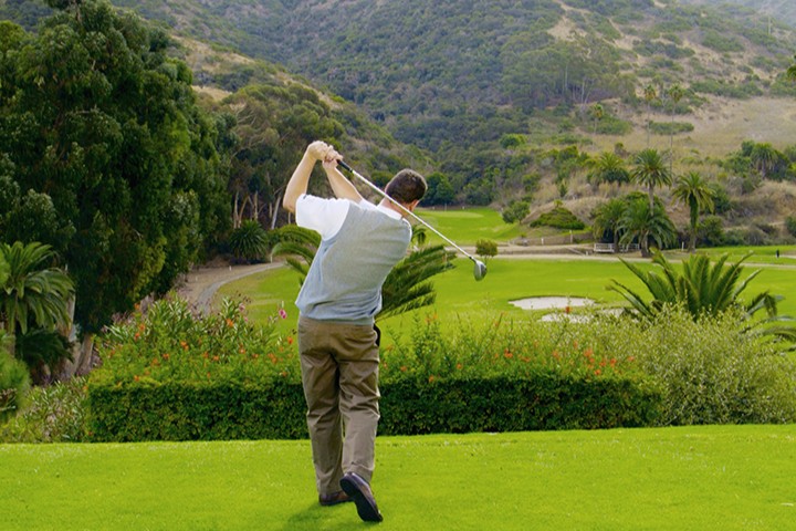 Golfer swings club on lush green course surrounded by trees and hills.
