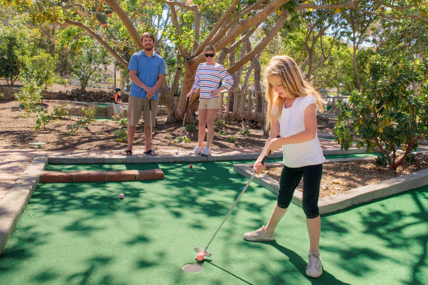 Young girl playing mini golf while two adults watch in a sunny park setting.