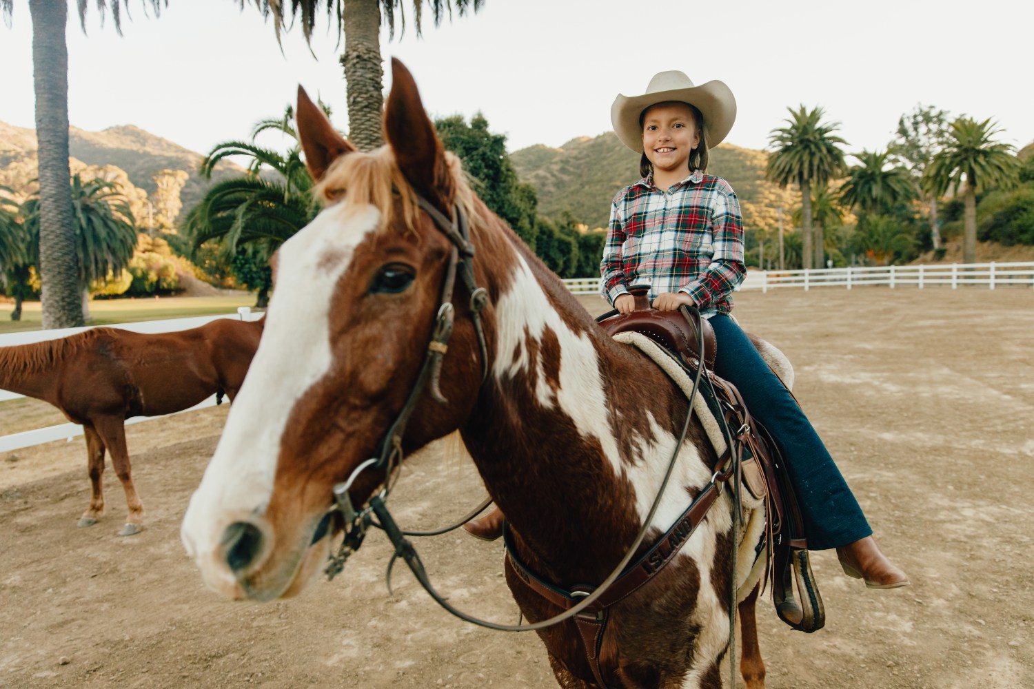 Child in plaid shirt and cowboy hat riding a brown and white horse in a fenced area with palm trees.