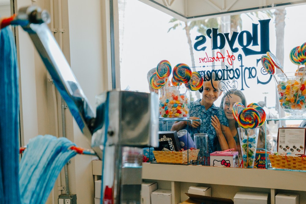 Two kids smiling through candy shop window with colorful lollipops and sweets.
