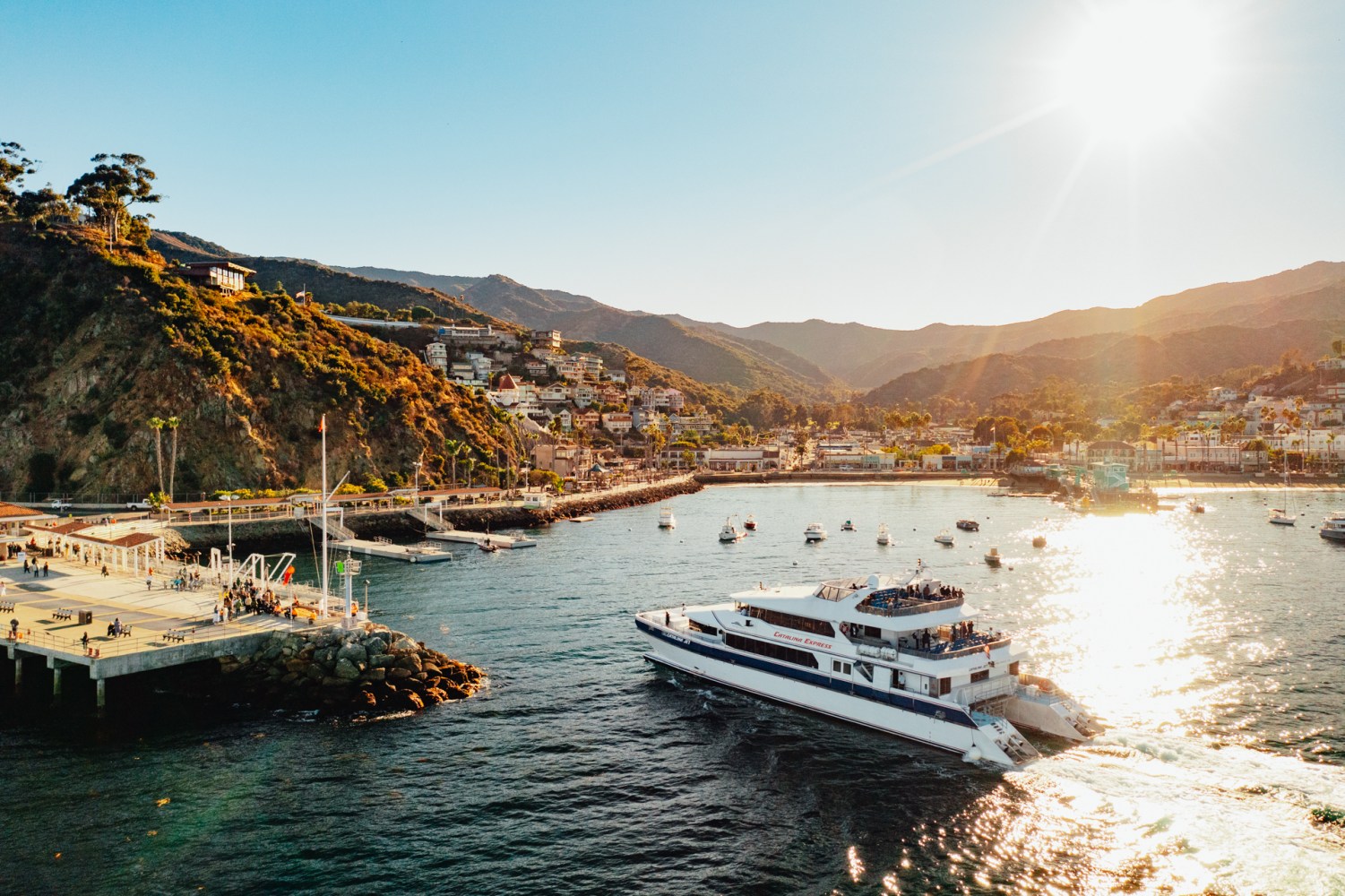 Ferry sailing near a coastal town with mountains and sunset in the background.