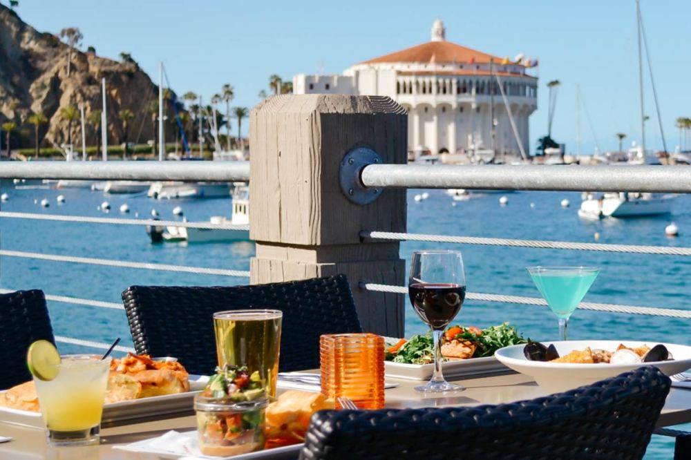 Outdoor dining table with food, drinks, and ocean view of a coastal building and boats.