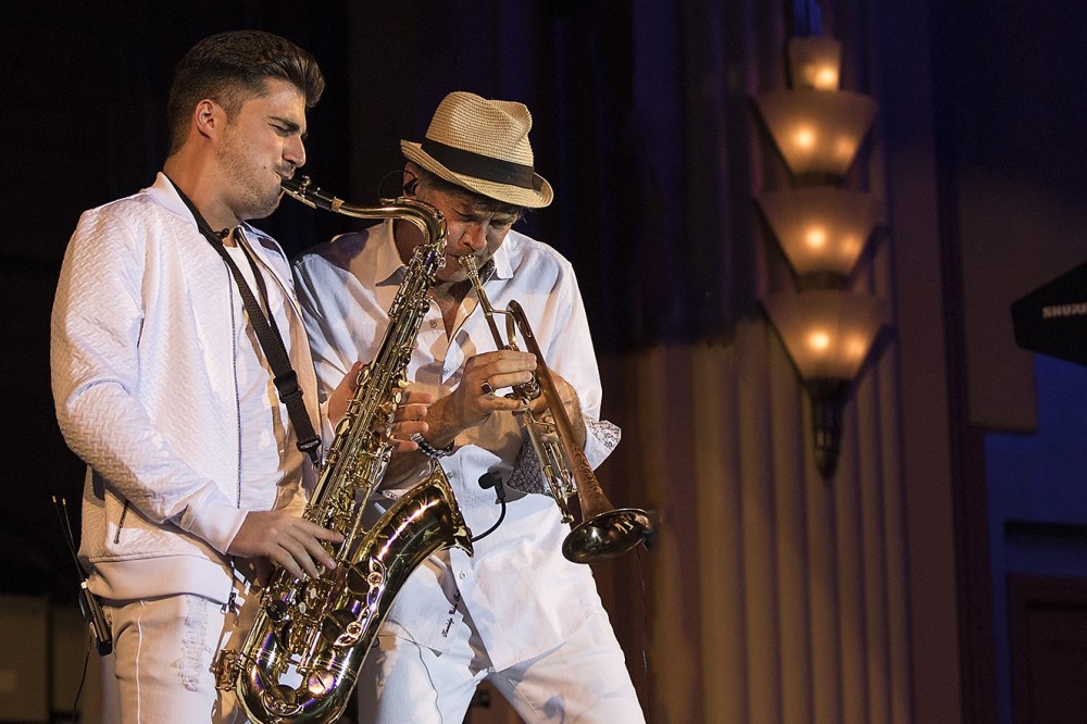 Two musicians in white outfits playing saxophone and trumpet on stage.